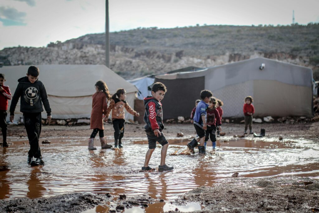 Kids in a refugee camp in Idlib, Syria, playing in muddy water, reflecting resilience and community spirit.