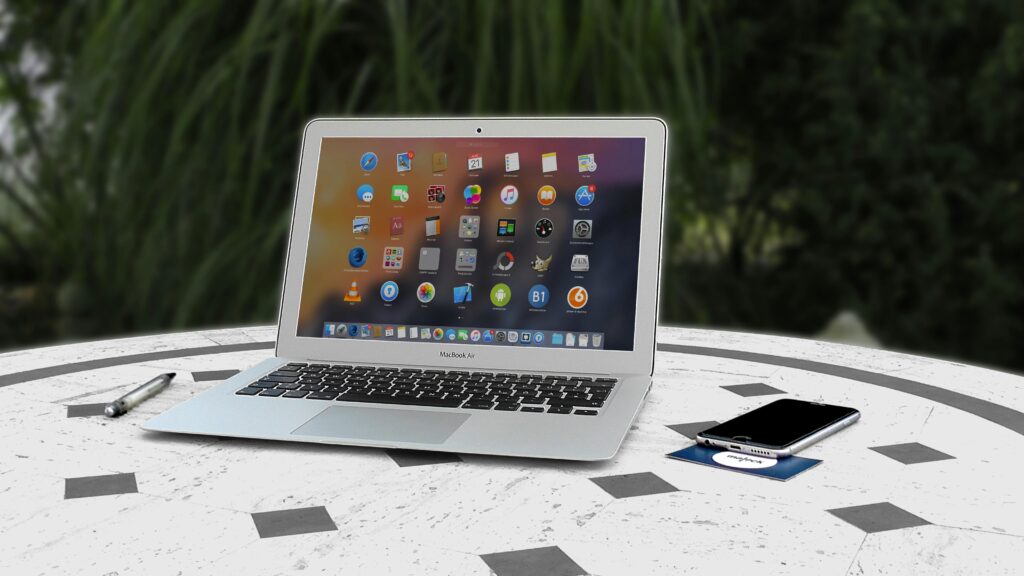 Open laptop with app icons displayed, smartphone and pen on white table outdoors.